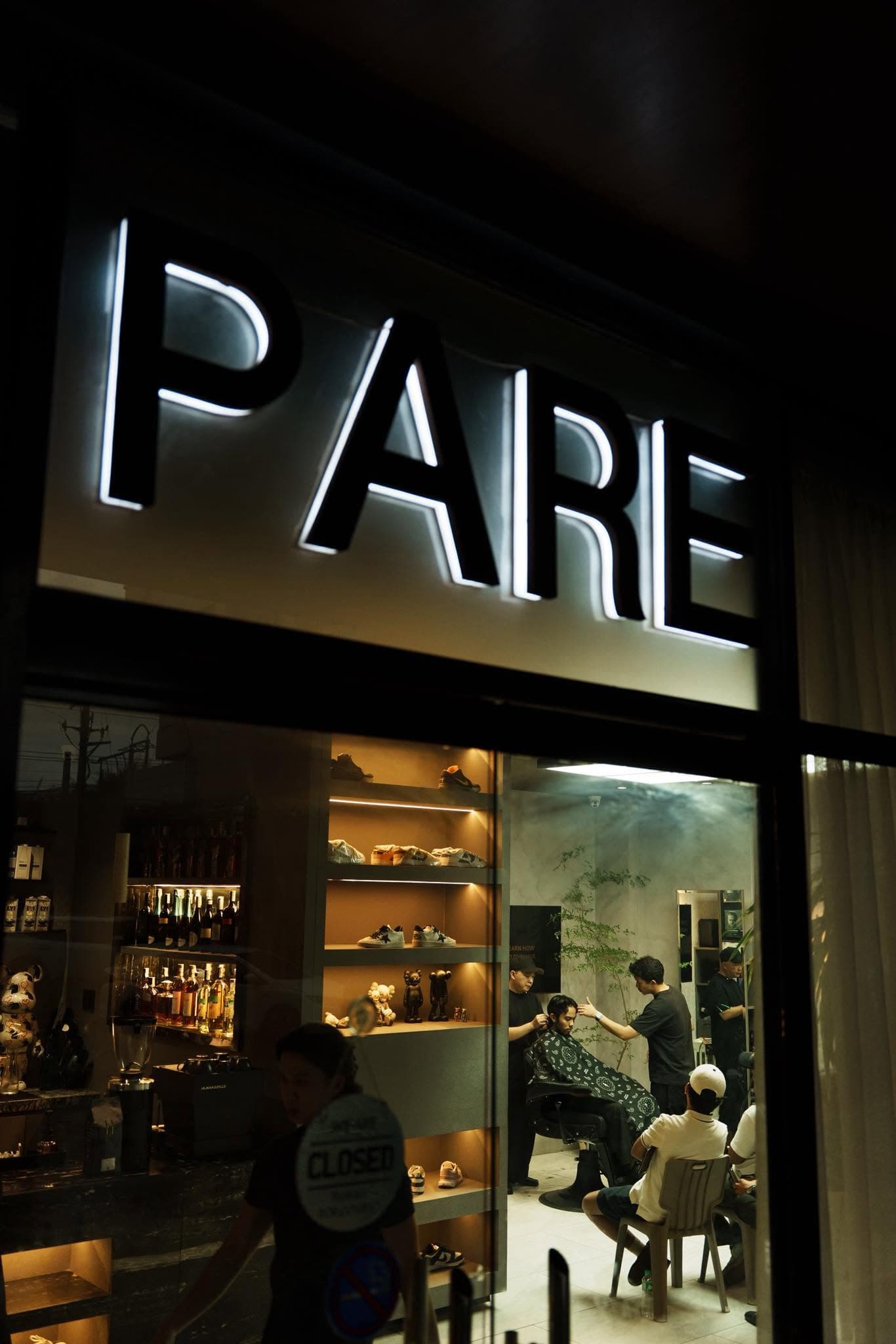 The PARE storefront at night, barbers at work inside